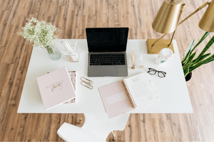 Image on a woman's wooden desk with a laptop, pink notebooks, laptop, flowers, glasses and lamps. Learn the best
blogging tips for beginners with this post 