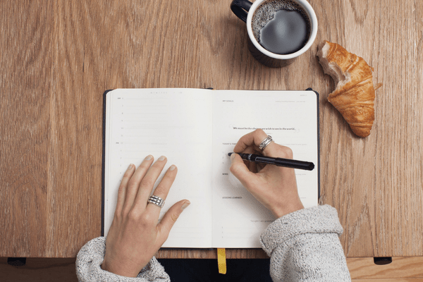 Woman writing a journal with a coffee and croissant on the table. This image is in relation to a blog post about bullet journal: What to Track in Your Habit Tracker