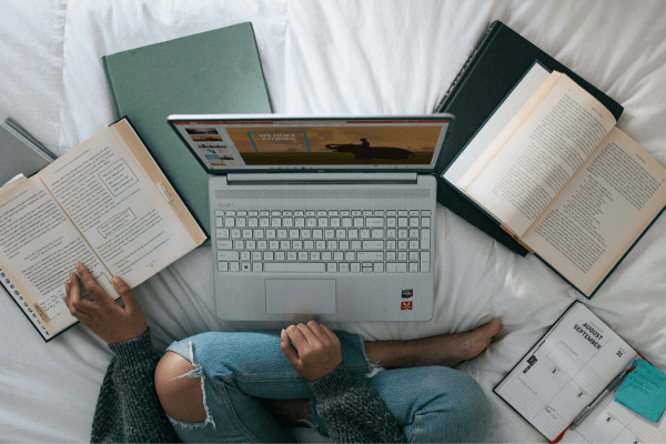 Image of a blogger (visible her only legs and hands) reading something on her laptop in the bed and some books are laying around. She is looking for the best free courses online for bloggers
