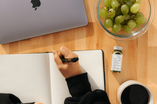 Image showing someone writing a journal, eating healthy (suggestion because of the fruits on the table). Blog post related to good daily habits to improve your life