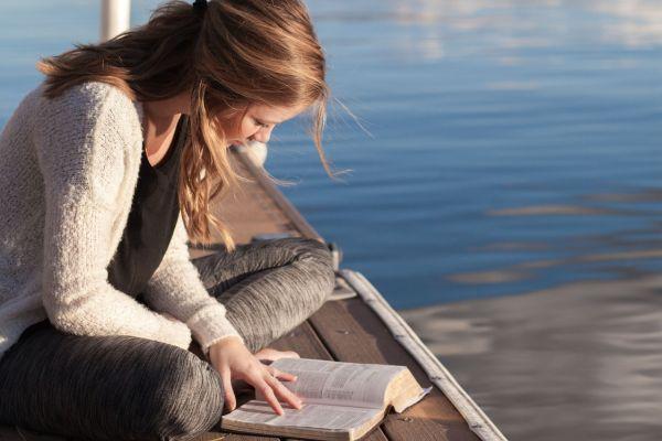a girl sitting next to a lake and reading a book. This photo is in relation to a blog post on best law of attraction books