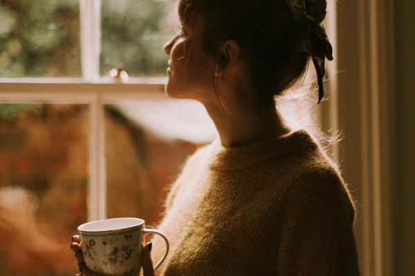 introverted woman looking trough the window with a cup of tea as a part of her daily self-care ritual.